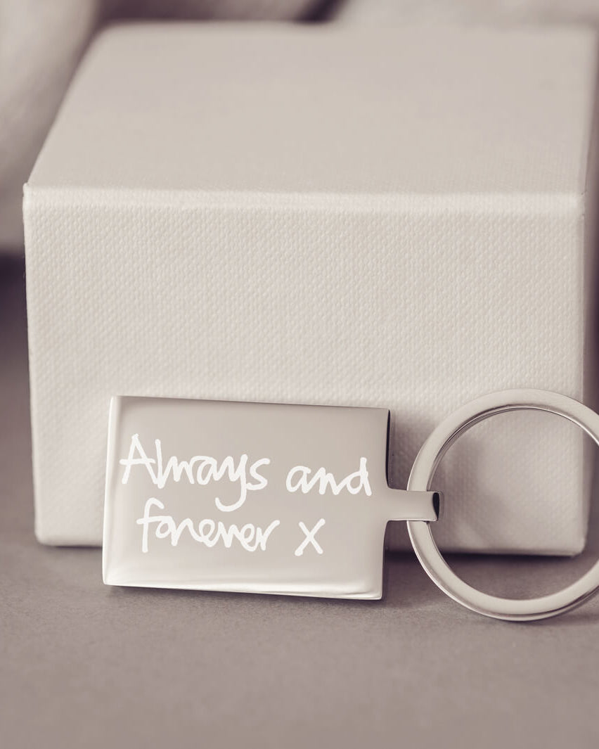 A stainless steel rectangular keyring, with a handwritten message of 'Always and forever x' engraved onto the front. Displayed against a light grey jewellery box.