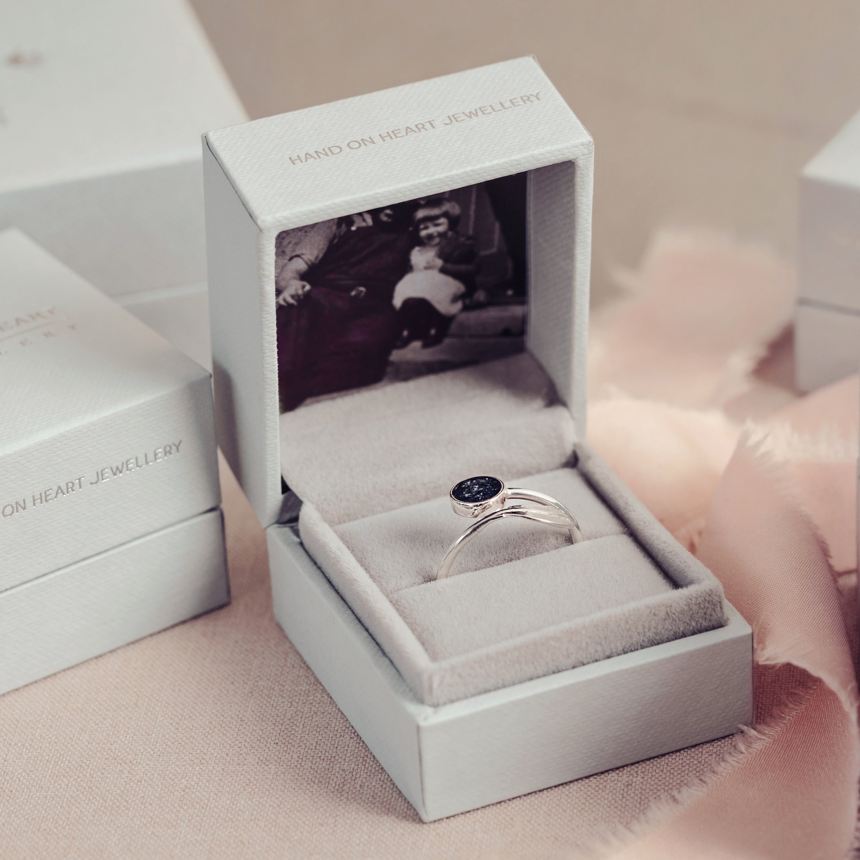 A silver ashes feather ring displayed in a soft grey jewellery box surrounded by similar jewellery boxes