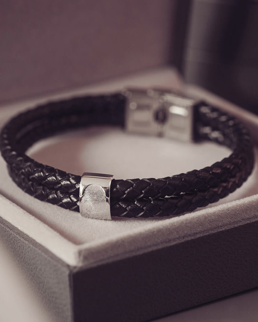 Black braided leather bracelet featuring a silver charm engraved with a fingerprint, displayed in a grey jewellery box
