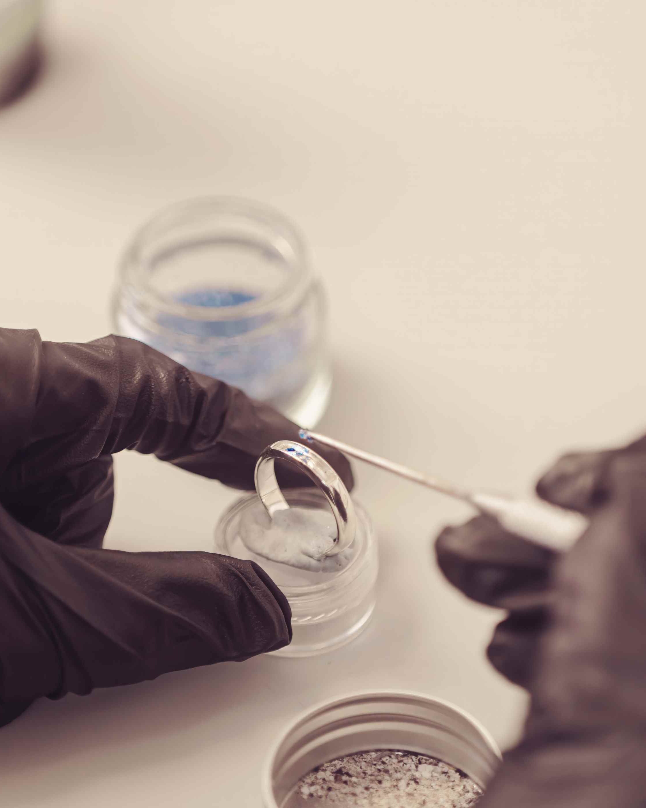 a close up of someone wearing black protective gloves creating a sterling silver womens ring band, carefully adding the ashes and blue crystal into the ring band using a pointed metal tool