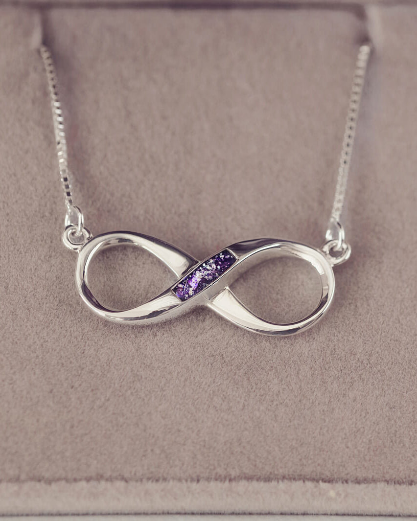 Sterling silver infinity necklace with a central section filled with memorial ashes and purple crushed crystals, displayed on a beige fabric background