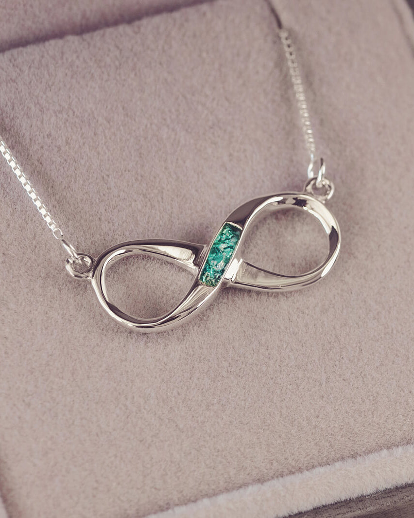 Sterling silver infinity necklace with a central section filled with memorial ashes and green crushed crystals, displayed on a beige fabric background