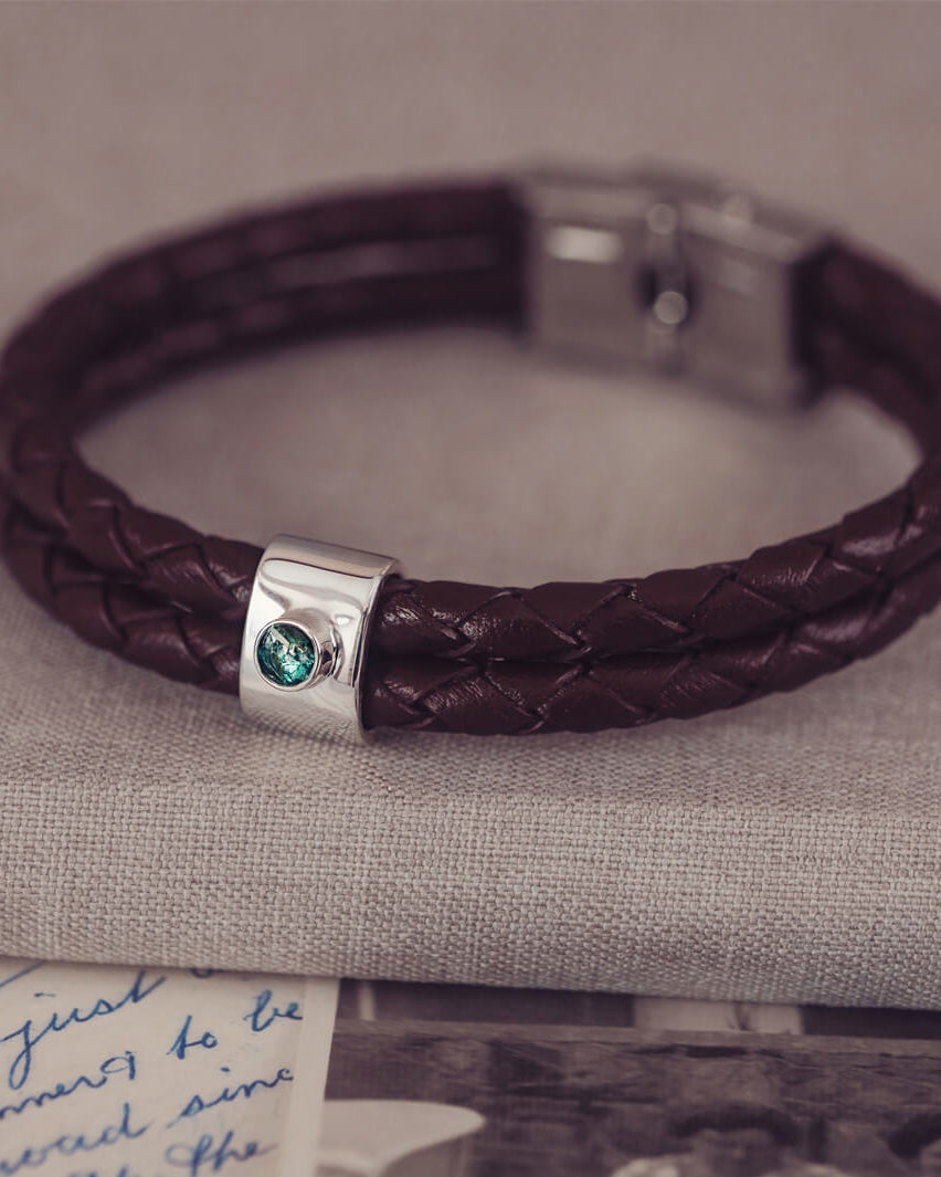 Brown braided leather bracelet featuring a silver charm with a small green memorial stone set in the centre, displayed on a grey surface.