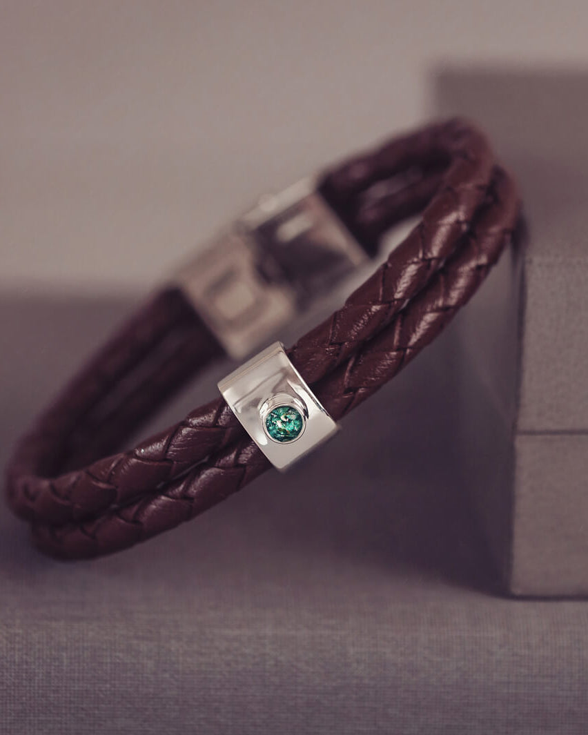 Brown braided leather bracelet featuring a silver charm with a small green memorial stone set in the centre, displayed on a grey surface.