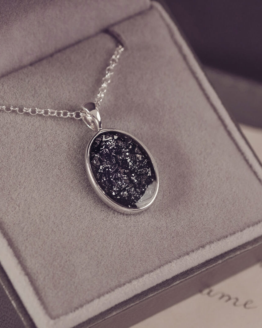 Sterling silver oval necklace with a central section filled with memorial ashes and black crushed crystals, displayed in a grey jewellery box