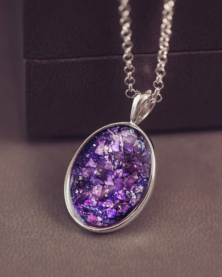 Sterling silver oval necklace with a central section filled with memorial ashes and purple crushed crystals, displayed on a grey jewellery box