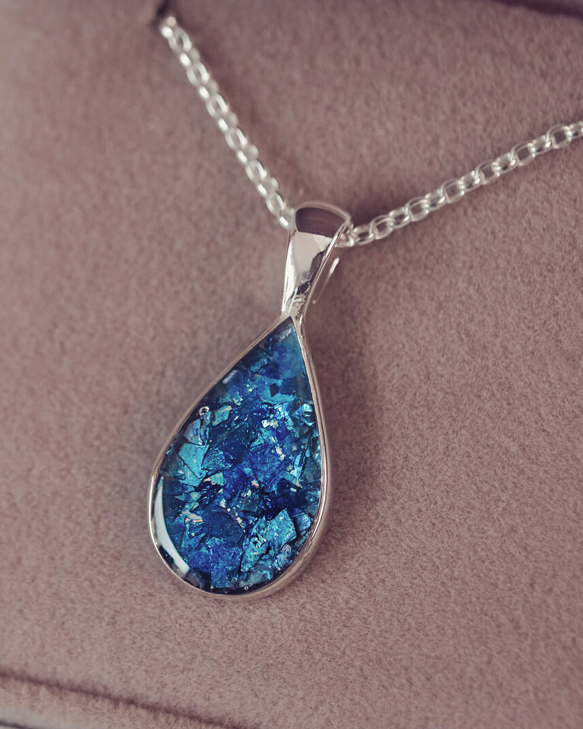 Sterling silver teardrop necklace with a central section filled with memorial ashes and blue crushed crystals, displayed on a grey fabric background