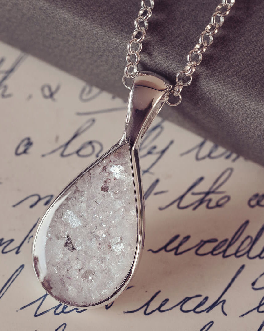 Sterling silver teardrop necklace with a central section filled with memorial ashes and white and clear crushed crystals, displayed on a white paper background with a handwritten message in blue ink