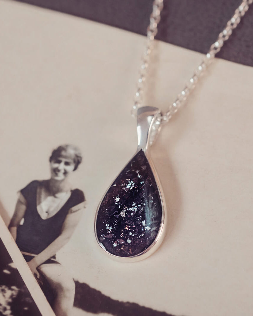 Sterling silver teardrop necklace with a central section filled with memorial ashes and black crushed crystals, displayed on a background of an old photograph of a woman in a swimming cap