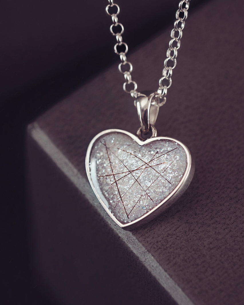 A silver heart shaped necklace charm, on a silver chain, filled with shite glitter, strands of dark hair, and resin.