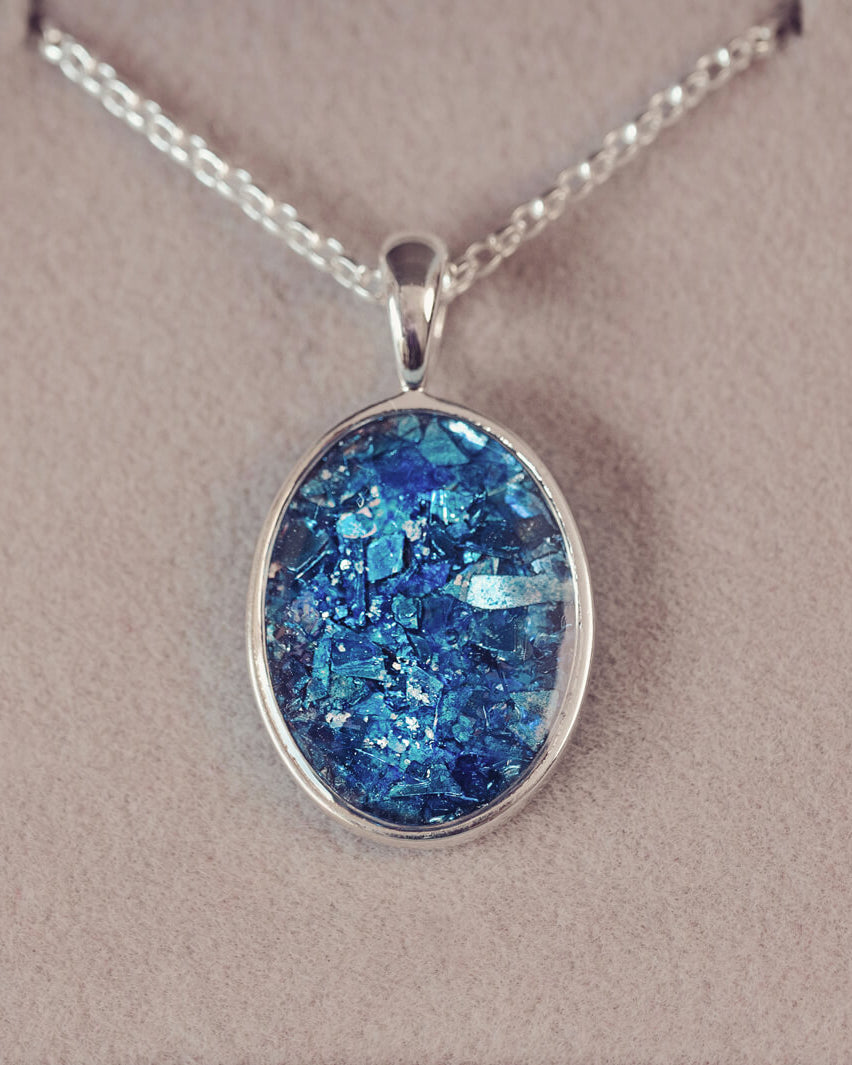 Sterling silver oval necklace with a central section filled with memorial ashes and blue crushed crystals, displayed on a grey fabric background