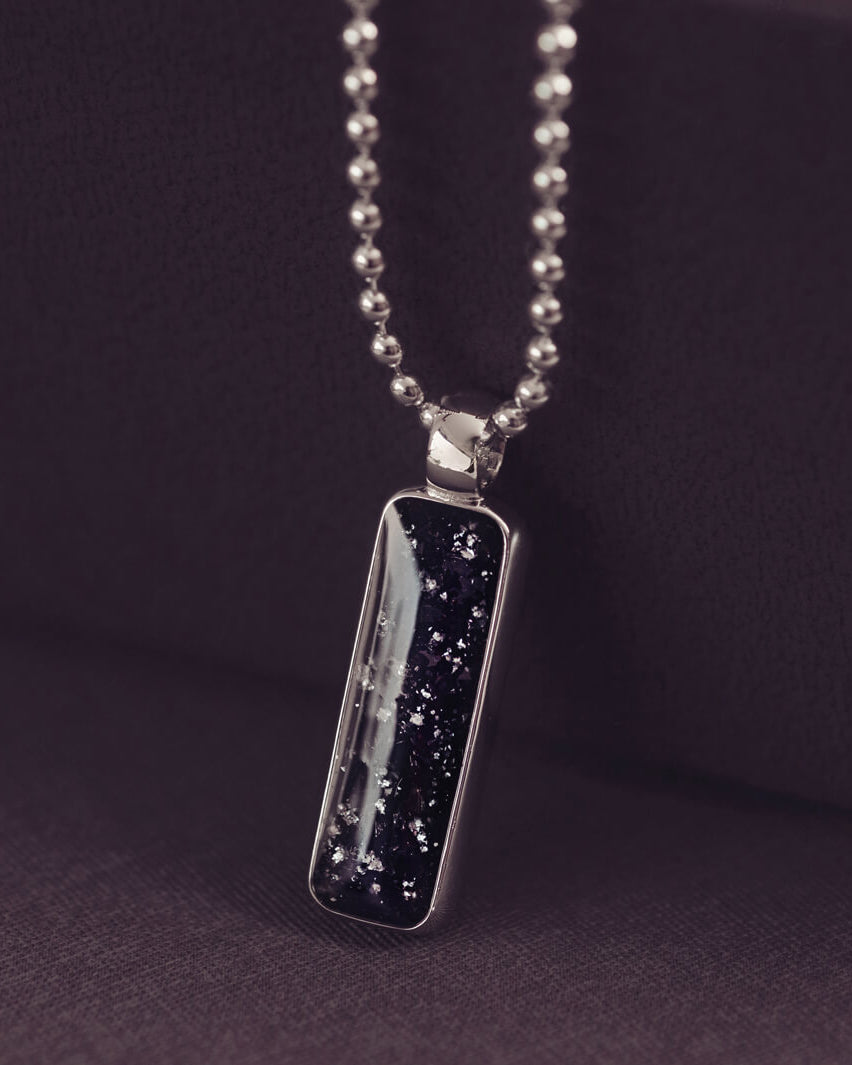 A sophisticated Mens silver necklace featuring a rectangular pendant filled with shimmering black crystal and resin, delicately layered with cremation ashes, showcased against a dark background on a silver ball chain
