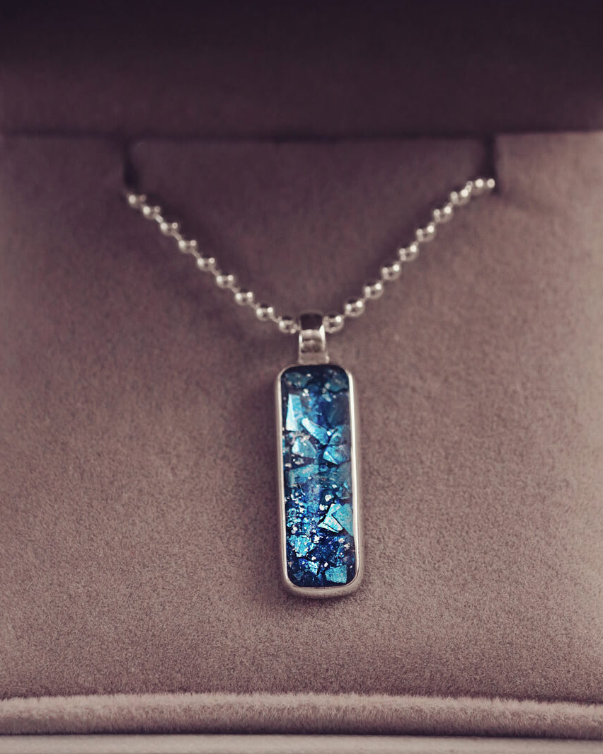A sophisticated Mens silver necklace featuring a rectangular pendant filled with shimmering blue crystal and resin, delicately layered with cremation ashes, showcased against a dark background on a silver ball chain