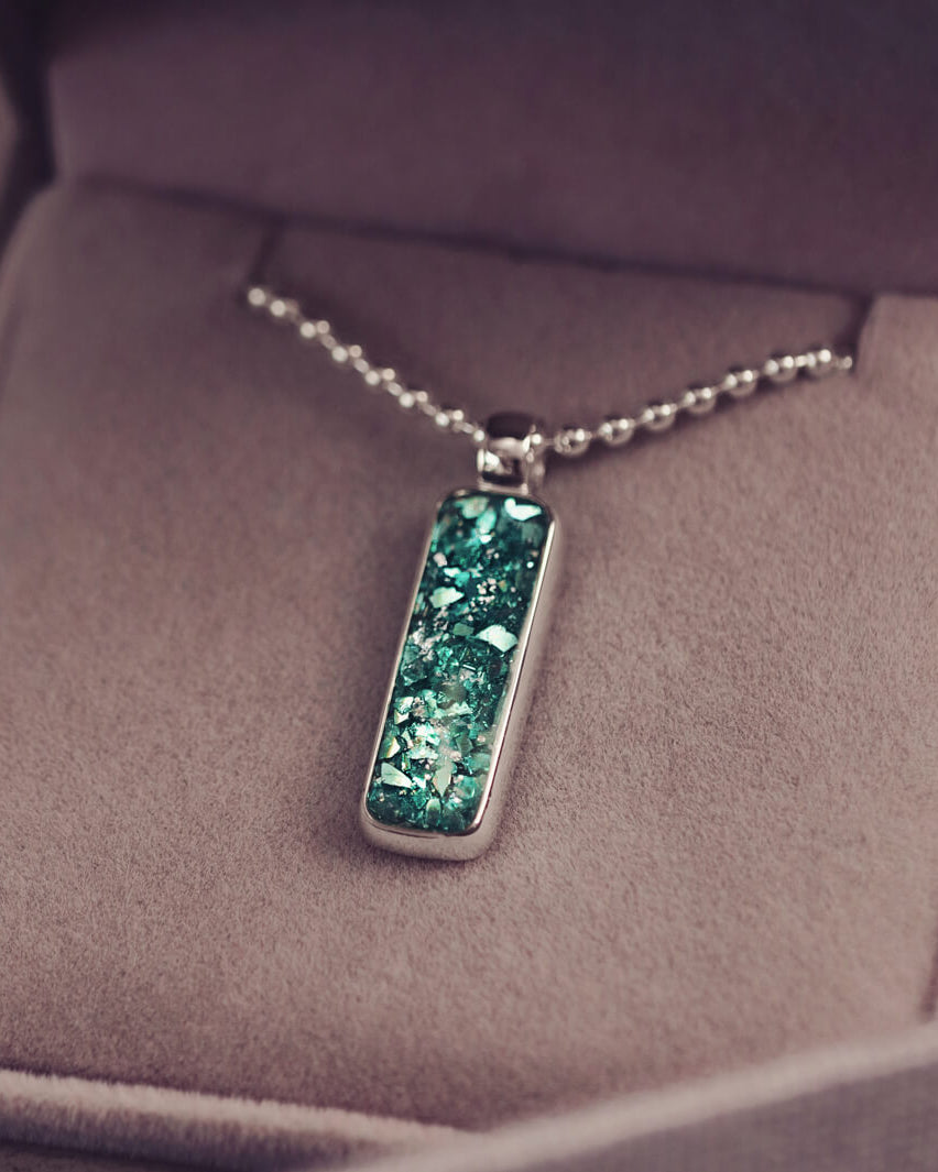 A sophisticated Mens silver necklace featuring a rectangular pendant filled with shimmering green crystal and resin, delicately layered with cremation ashes, showcased against a dark background on a silver ball chain