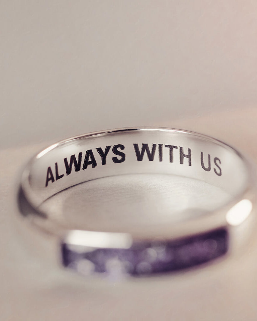 Sterling silver Mens Band Ring with a central section filled with memorial ashes and purple crushed crystals, displayed on a blurred pale pink background. The ring has the message  'ALWAYS WITH US' engraved into the inside of the band in black