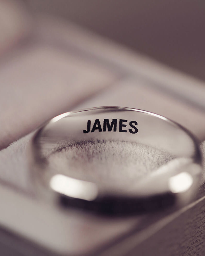 Sterling silver Mens Band Ring with a central section filled with memorial ashes and black crushed crystals, displayed on a blurred grey background. The ring has the message 'JAMES' engraved into the inside of the ring band in black