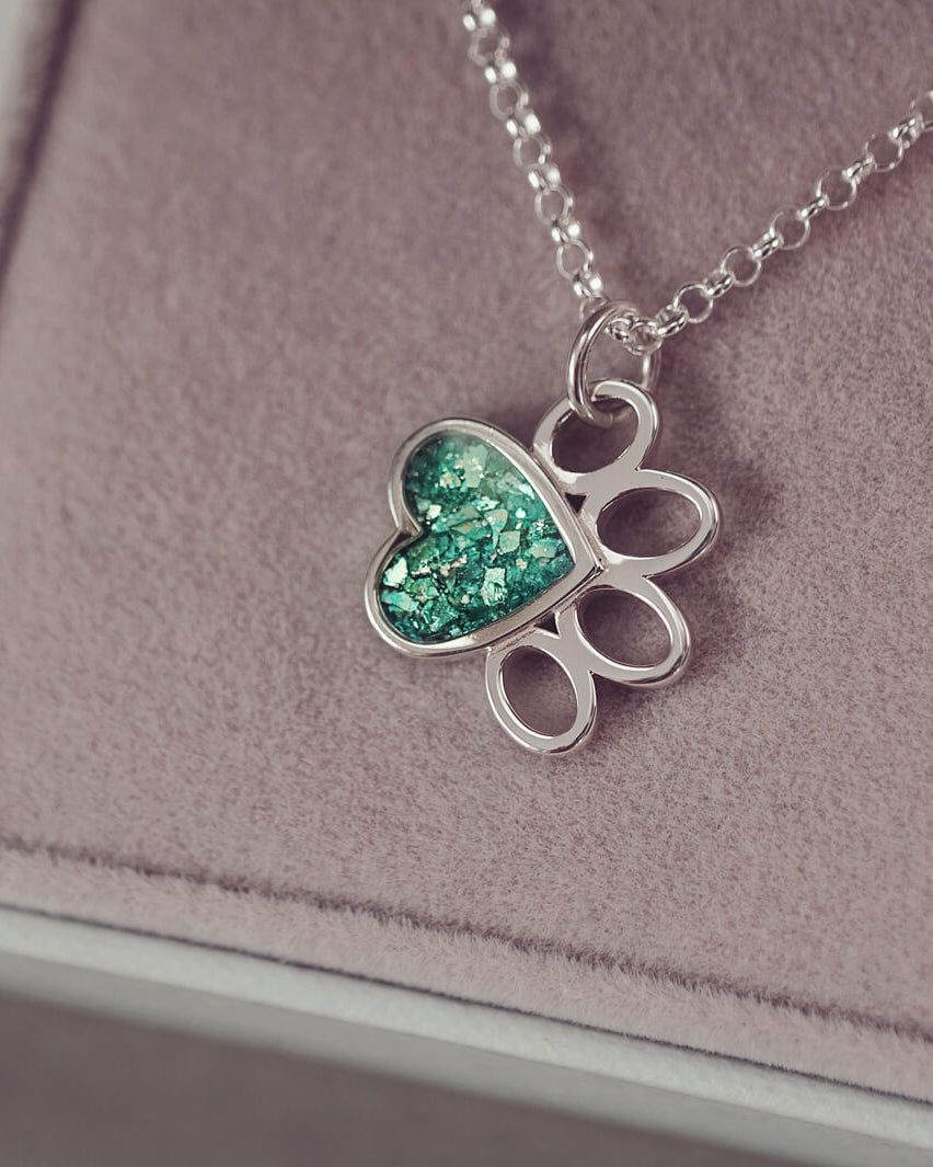 A Pawprint Shaped Sterling Silver Necklace, where the main pad is filled with green crystal and cremation ashes. Hanging from a sterling silver belcher chain in a grey jewellery box.