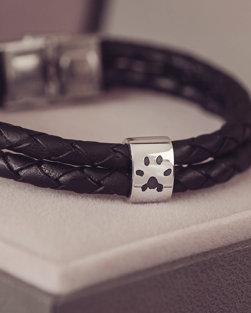 Black braided leather bracelet featuring a silver charm engraved with a black pawprint, displayed on a soft grey background.