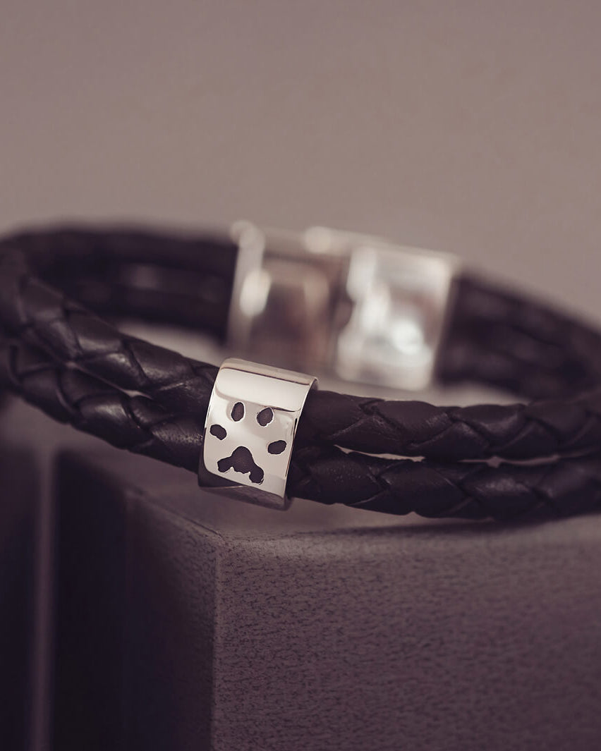 Black braided leather bracelet featuring a silver charm engraved with a black pawprint, displayed on a soft grey background.