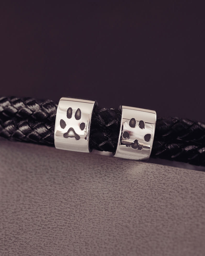 Black braided leather bracelet featuring two sterling silver charms, each engraved with a black pawprint, displayed on a soft grey background.