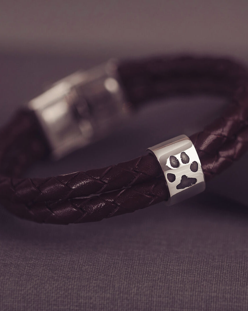 Brown braided leather bracelet featuring a silver charm engraved with a black pawprint, displayed on a dark grey background.