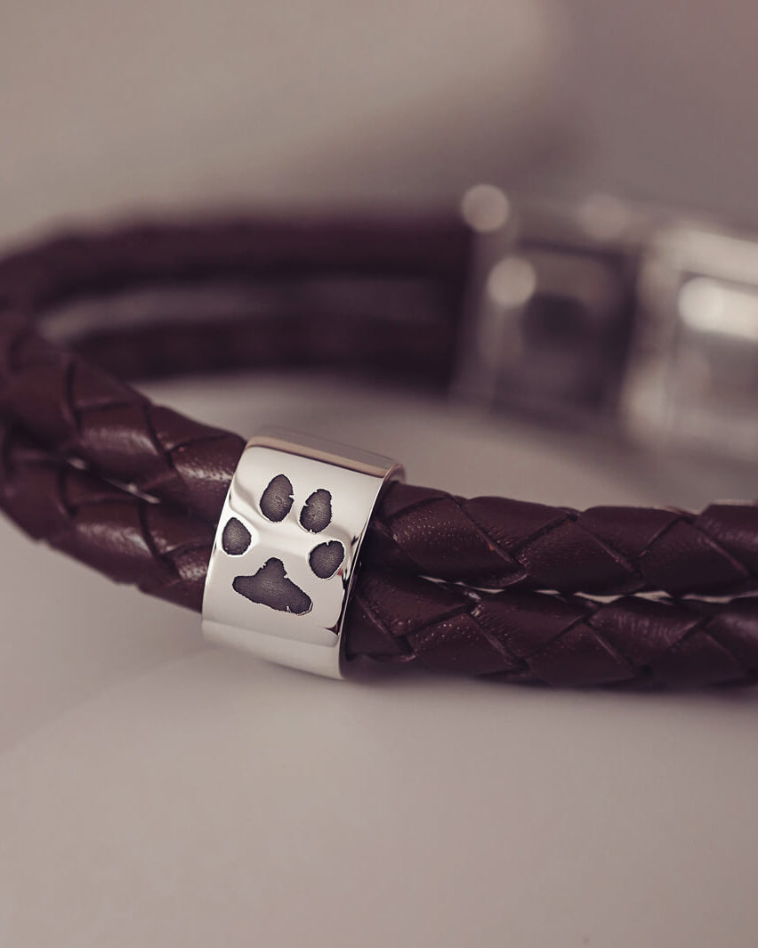 Brown braided leather bracelet featuring a silver charm engraved with a black pawprint, displayed on a soft grey background.