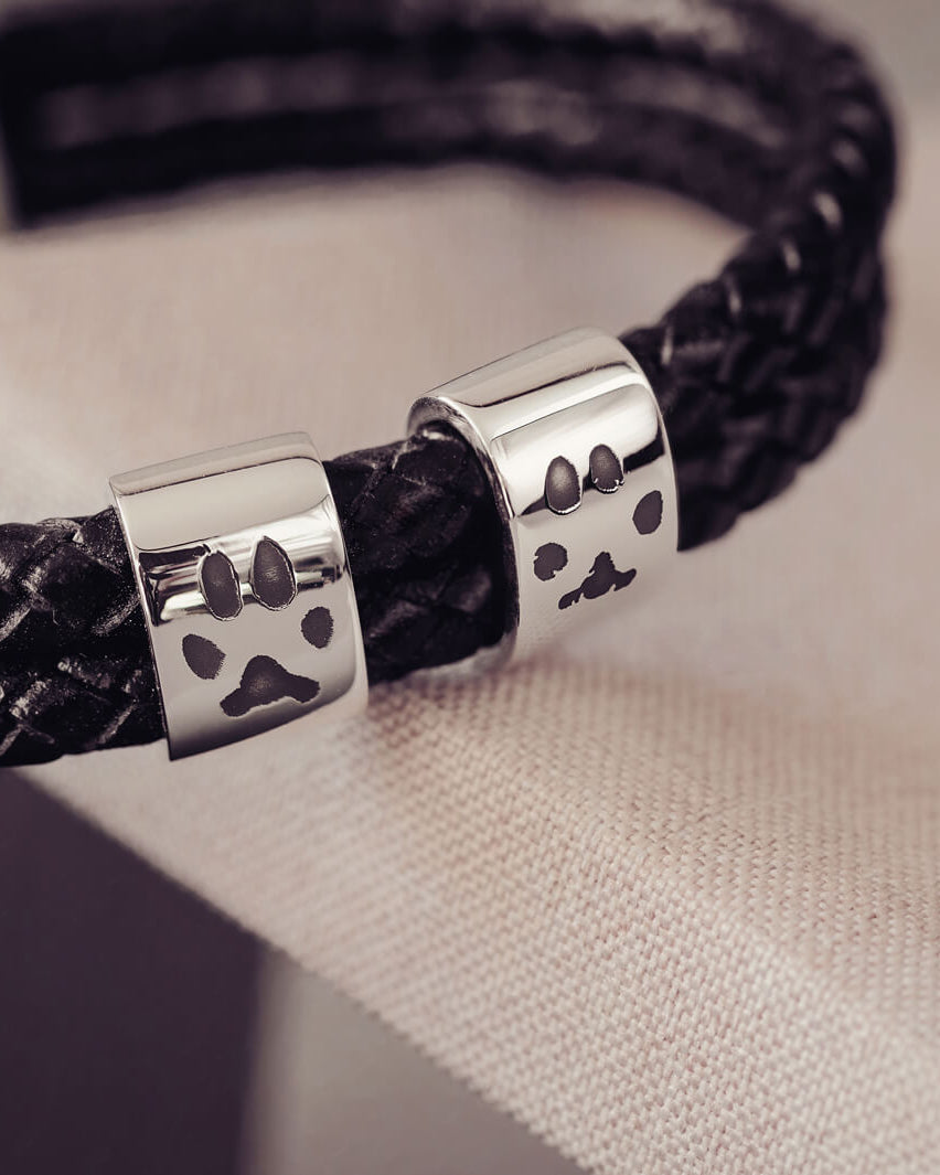 Black braided leather bracelet featuring two sterling silver charms, each engraved with a black pawprint, displayed on a soft grey background.