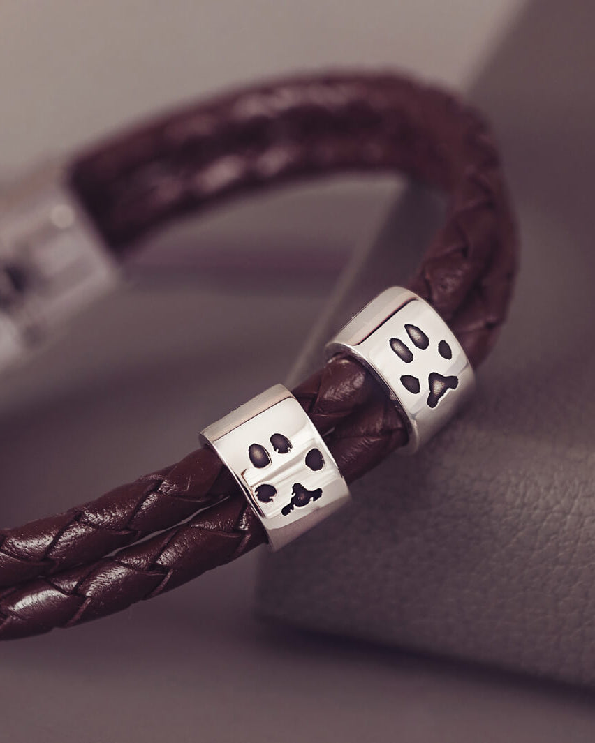 Brown braided leather bracelet featuring two sterling silver charms, each engraved with a black pawprint, displayed on a soft grey background.