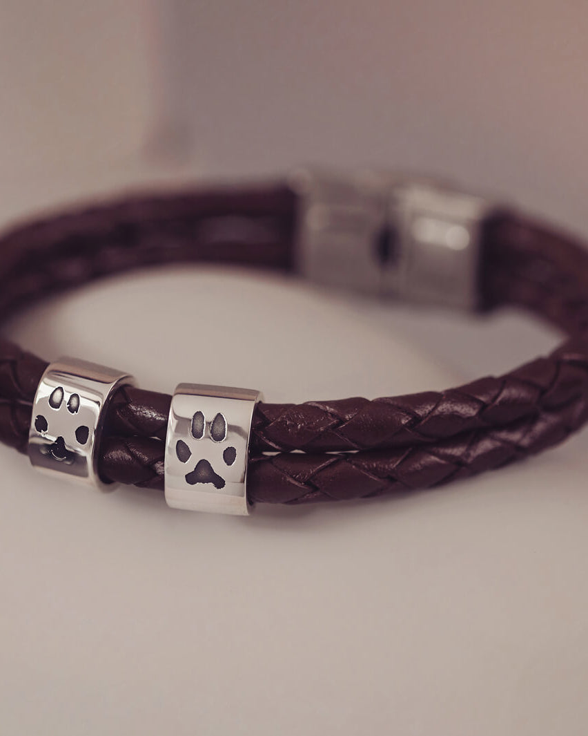 Black braided leather bracelet featuring two sterling silver charms, each engraved with a black pawprint, displayed on a soft grey background.