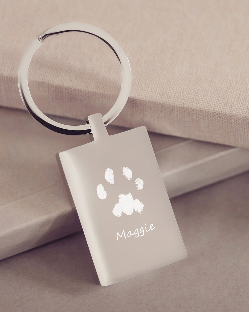 A rectangular stainless steel keyring, engraved with one pawprint and name, leaning on a dusky pink fabric background