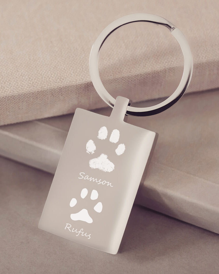 A rectangular stainless steel keyring, engraved with two pawprints and names, leaning on a dusky pink fabric background