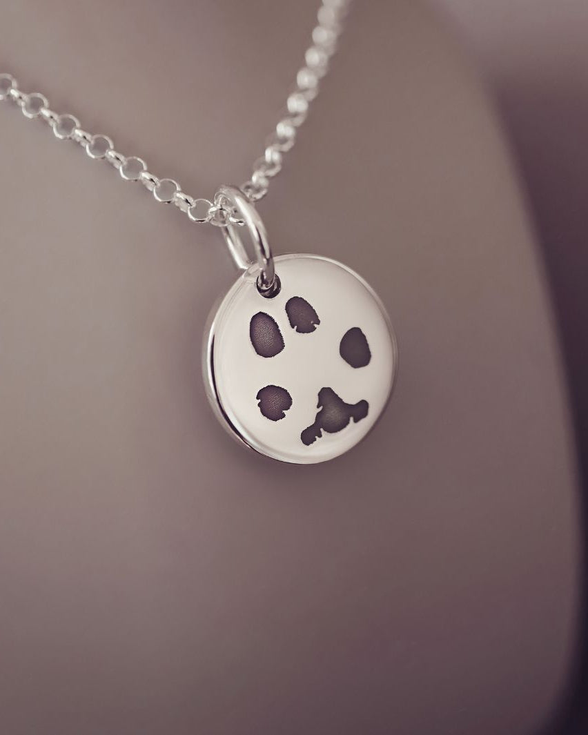 Round sterling silver charm on a silver chain, the charm features an imprinted pawprint in black. The necklace is on a pinky-grey background