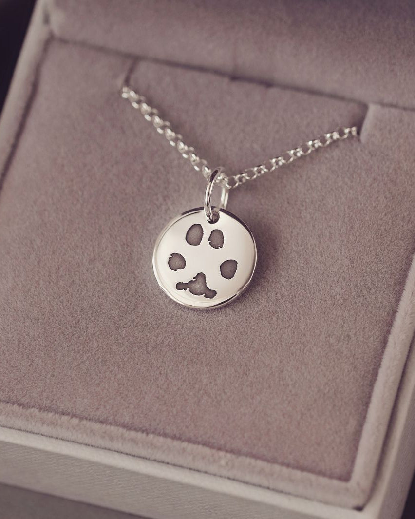 Round sterling silver charm on a silver chain, the charm features an imprinted pawprint in black. The necklace is in a grey jewellery box.