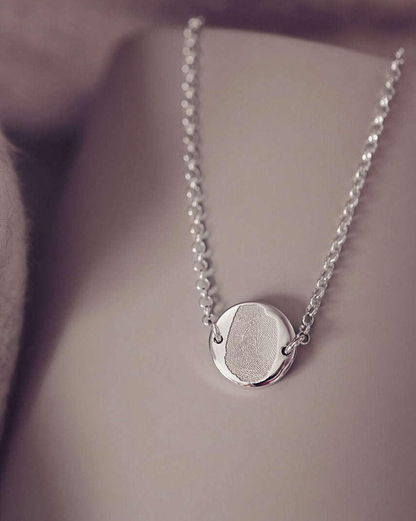 A round silver pendant bracelet featuring a detailed imprinted fingerprint, displayed on a delicate silver chain. The necklace rests in an elegant, soft velvet jewellery box