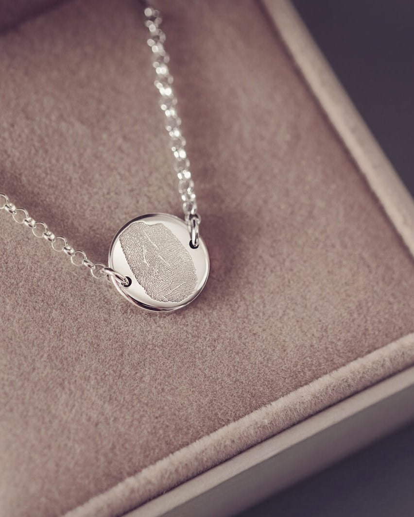 A round silver pendant bracelet featuring a detailed imprinted fingerprint, displayed on a delicate silver chain. The necklace rests in an elegant, soft velvet jewellery box