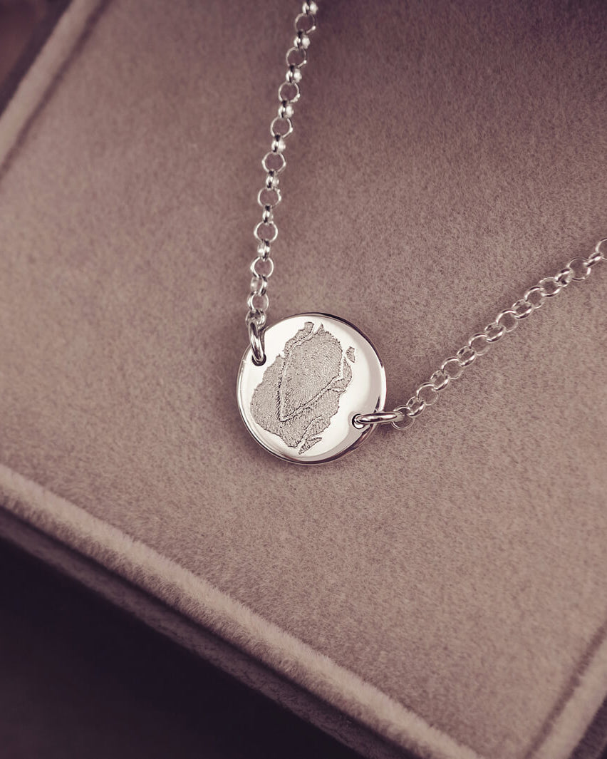 A round silver pendant bracelet featuring a detailed imprinted fingerprint, displayed on a delicate silver chain. The necklace rests in an elegant, soft velvet jewellery box