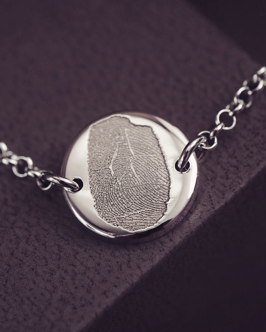 A round silver pendant bracelet featuring a detailed imprinted fingerprint, displayed on a delicate silver chain. The necklace rests in an elegant, soft velvet jewellery box