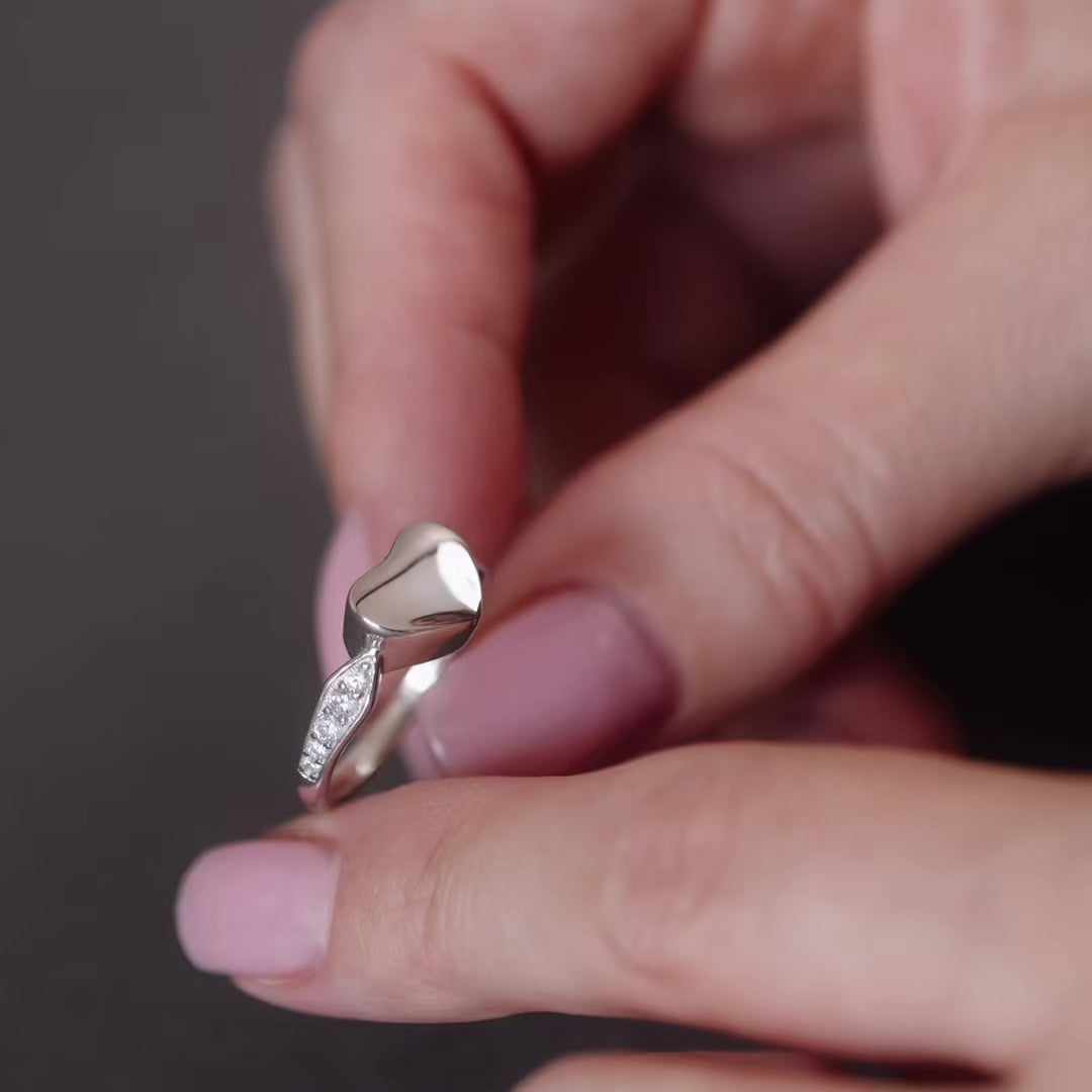Demo video of a heart ashes urn ring, it is silver with gems on the sides of the band. The video shows a women's hand holding the ring then unscrewing the top of the ring to then fill it with ashes.