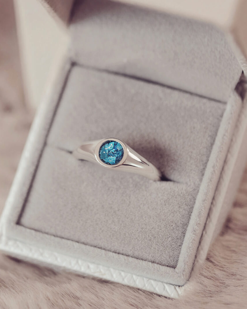 sterling silver ring band with circular stone holding cremation ashes and blue crystal sat in a grey jewellery box