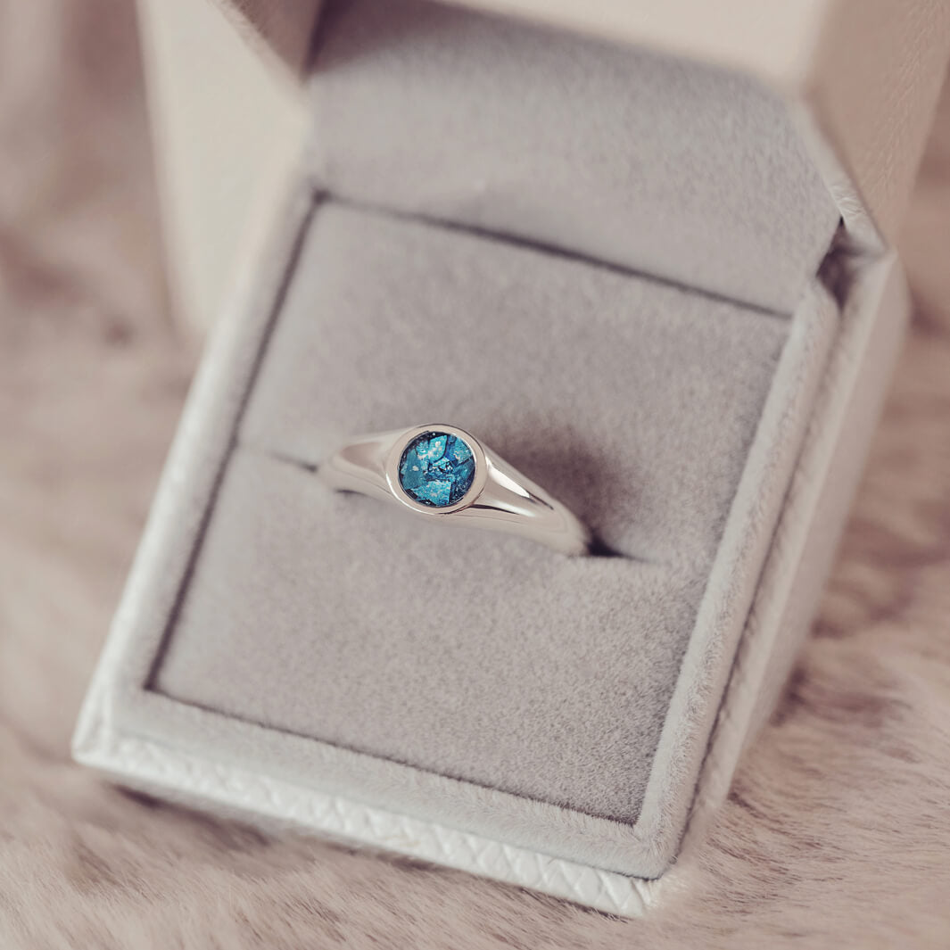 sterling silver ring band with circular stone holding cremation ashes and blue crystal sat in a grey jewellery box