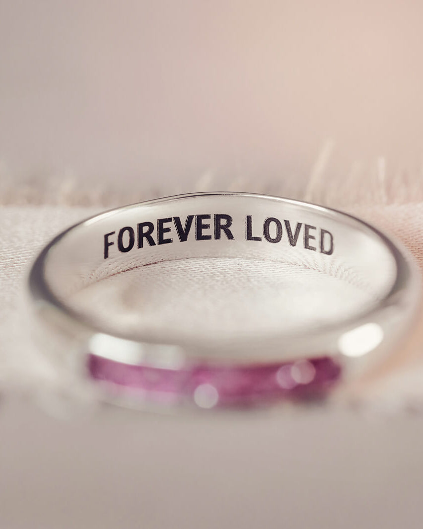 Sterling silver womens ring band with a central section filled with  memorial ashes and pink crushed crystals, displayed on a blurred fabric background. The ring has the message 'Forever Loved' engraved into the inside of the ring band, in black