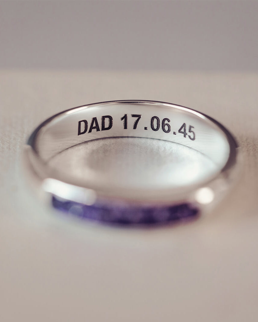 Sterling silver womens ring band with a central section filled with  memorial ashes and purple crushed crystals, on a blurred pale background. The ring has the message 'DAD 17.06.45' engraved into the inside of the ring band in black