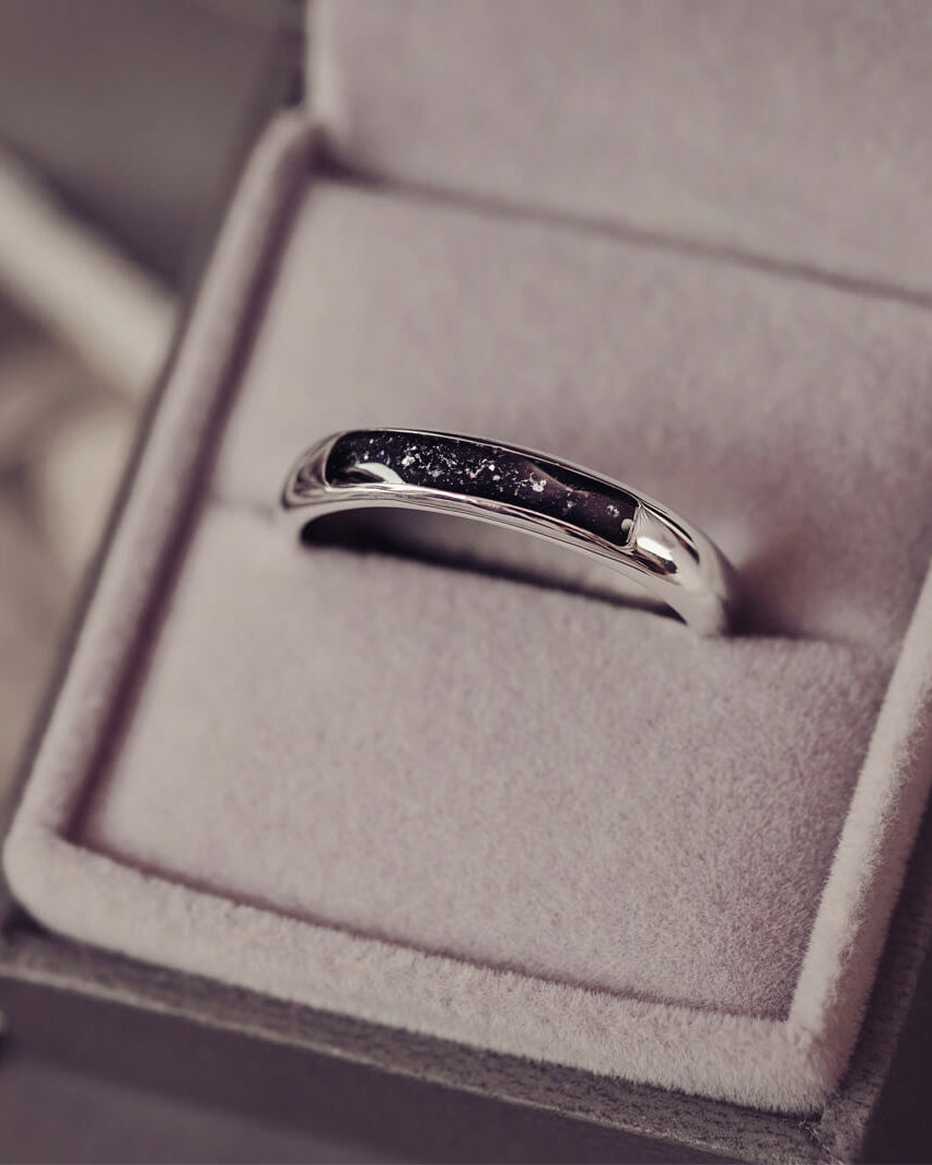 Sterling silver womens ring band with a central section filled with  memorial ashes and black crushed crystals, displayed in a grey jewellery box