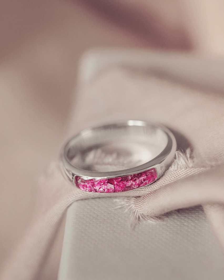 Sterling silver womens ring band with a central section filled with  memorial ashes and pink crushed crystals, displayed on a pink and grey fabric background