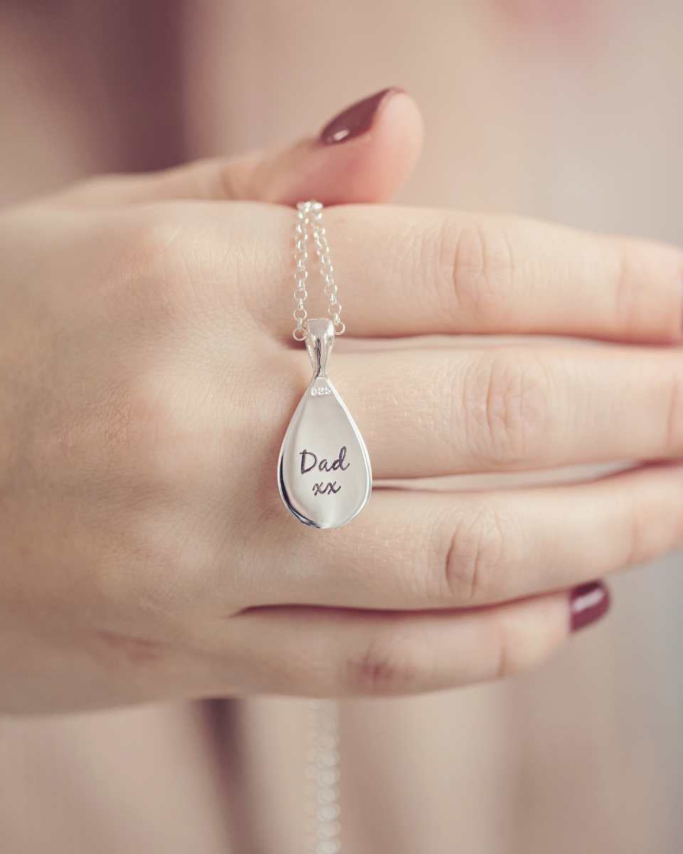 The reverse of a sterling silver teardrop shaped necklace, being held by a ladies hand with red painted nails. There is a message engraved into the back of the pendant which reads 'Dad xx'