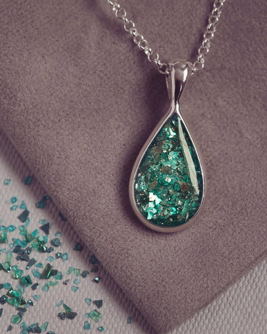 Sterling silver teardrop necklace with a central section filled with memorial ashes and green crushed crystals, displayed on a grey fabric background scattered with green crystal