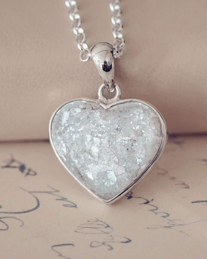 Sterling silver heart necklace with a central section filled with memorial ashes and white & clear crushed crystals, displayed on a beige fabric background.