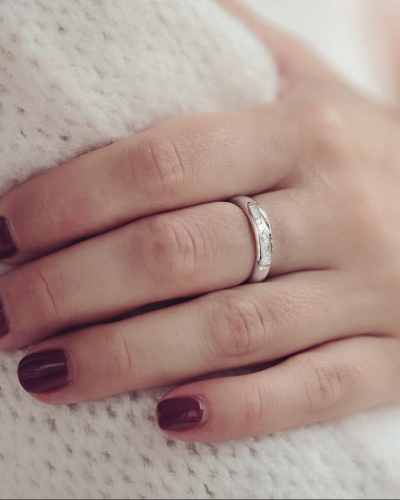 Close up of a ladies hand with red painted nails in a white jumper wearing Womens Cremation Ashes Ring Band, the middle of the ring band is filled with clear resin, ashes and white crushed crystals