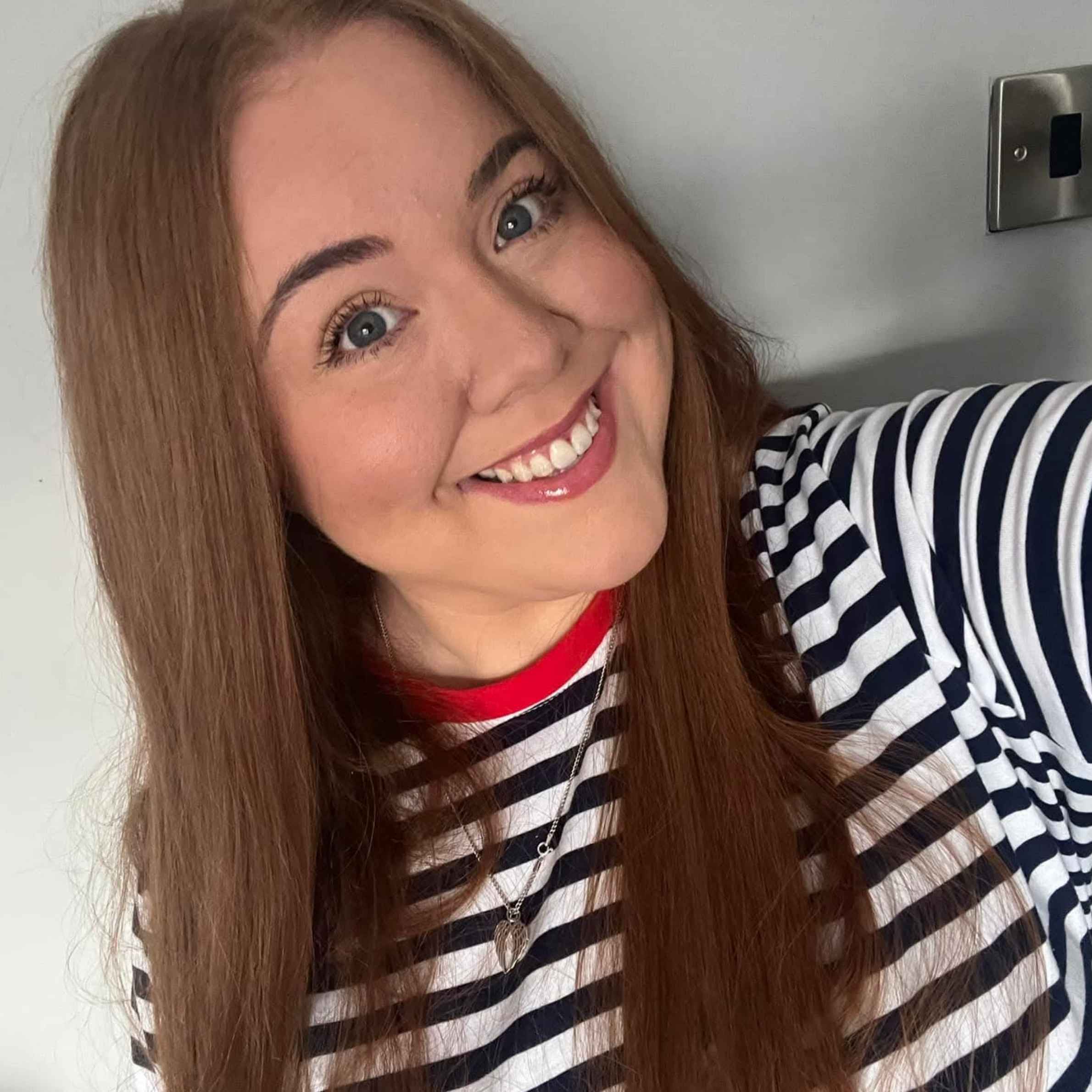 Smiling woman with long brown hair and blue eyes poses for a selfie indoors, wearing a navy-and-white striped shirt with red trim.