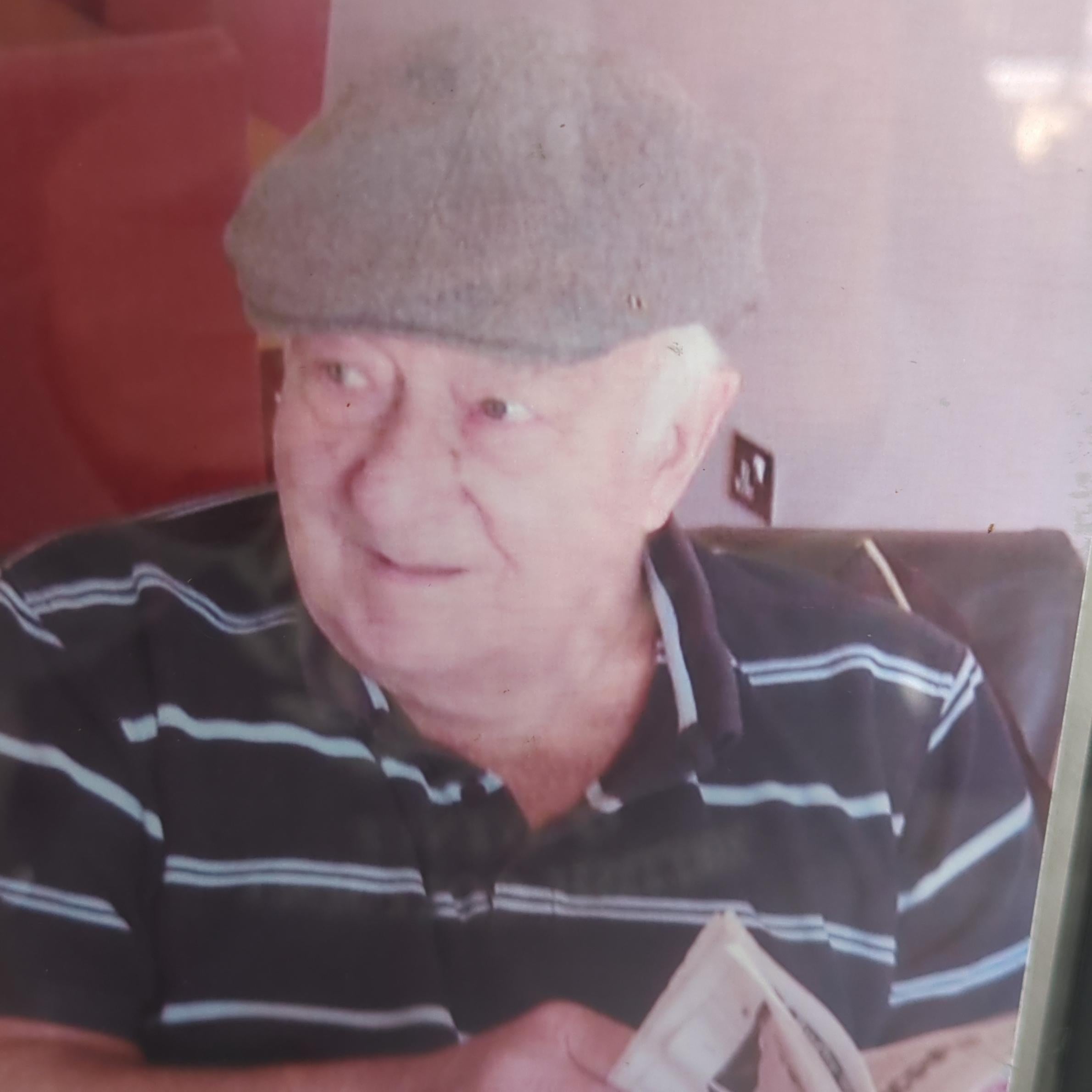 Man wearing navy blue polo shirt with horizontal baby blue stripes. He is wearing a grey flat cap and reading a newspaper.